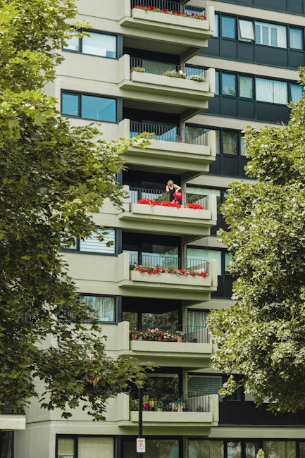 A multi-storey residential building with several concrete balconies featuring metal railings, some decorated with red and pink flower boxes. The building has large glass windows and a modern architect